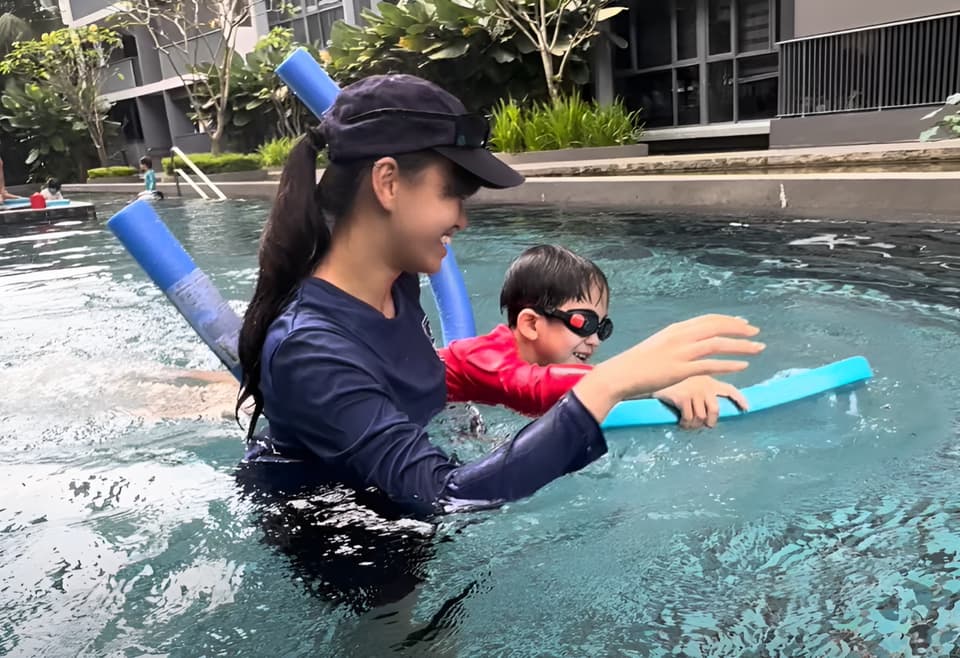 A smiling Poolva coach helping a boy in goggles kick with a float in a condo pool